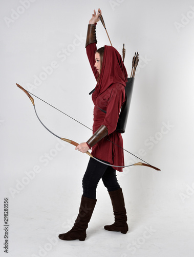 full length portrait of a brunette girl wearing a red fantasy tunic with hood, holding a bow and arrow. Standing pose on a white studio background.