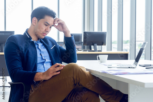 A young businessman sitting in a modern office. He has a feel stressed about the result of business profits not positive. On his table have a computer laptop tablet pen paper graph.