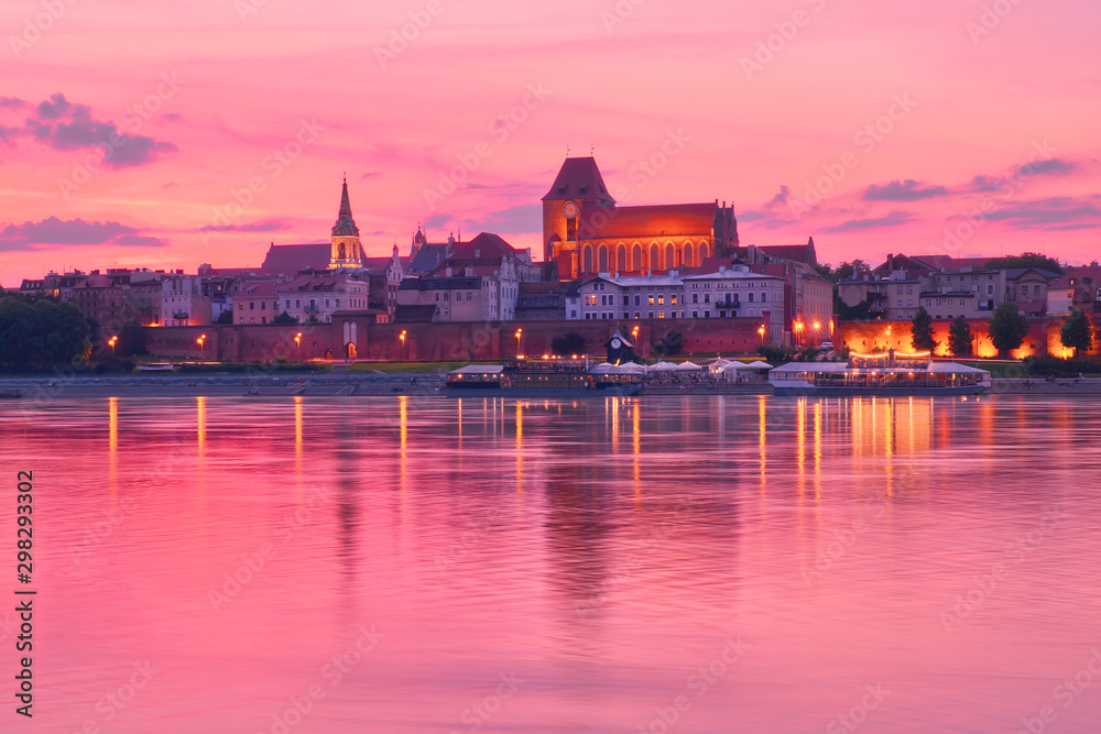 Fototapeta premium Toruńska starówka w Polsce, wpisana na Listę Światowego Dziedzictwa UNESCO, z iluminacją, odbita w Wiśle na różowym zachodzie słońca.
