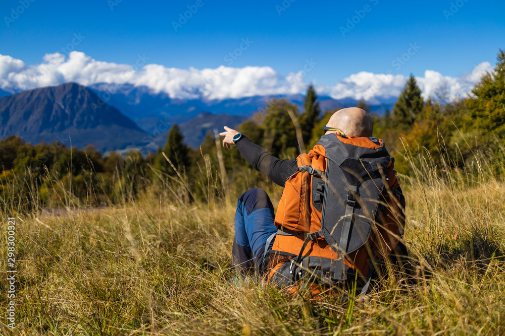 A male traveler with a backpack sits on the hill of a beautiful Alpine landscape and shows the direction with his hand