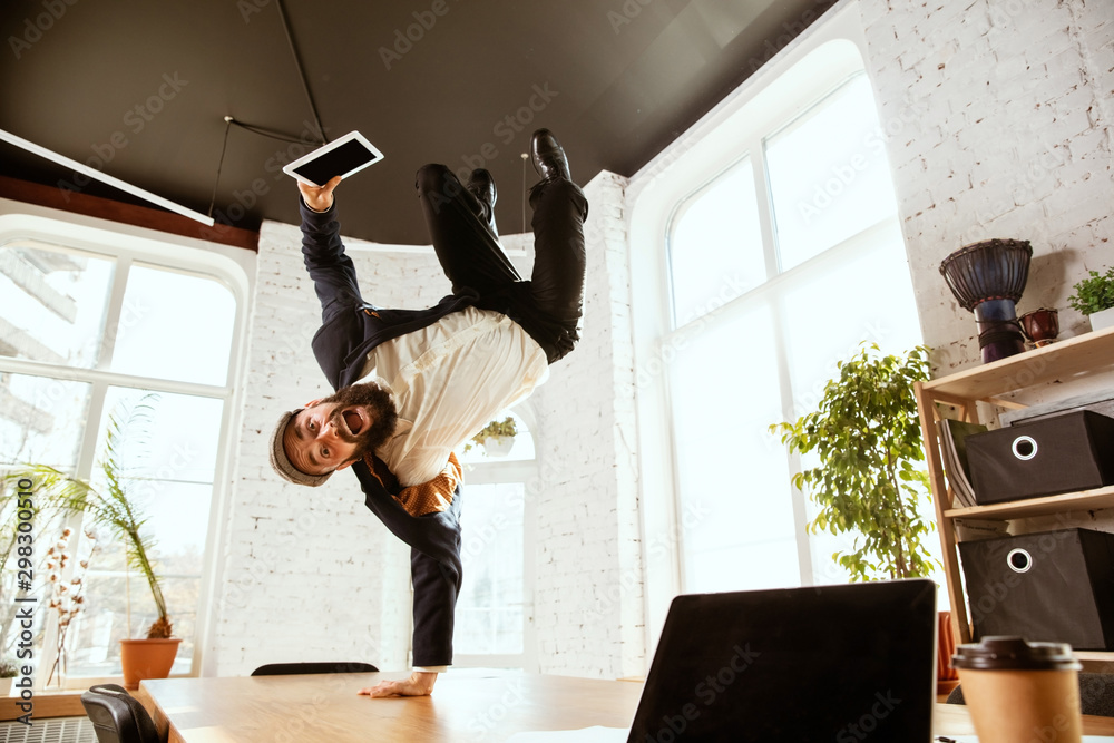 Fototapeta premium Young caucasian businessman having fun dancing break dance in the modern office at work time with gadgets. Management, freedom, professional occupation, alternative way of working. Loves his job.