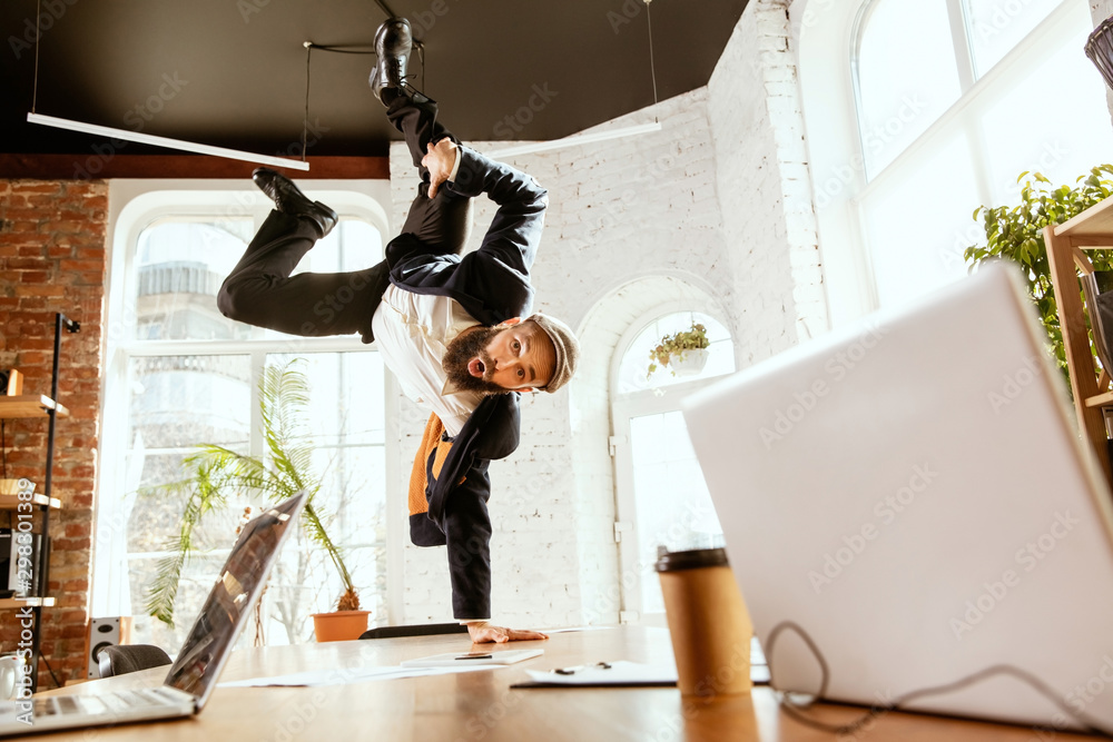 Young caucasian businessman having fun dancing break dance in the modern office at work time with gadgets. Management, freedom, professional occupation, alternative way of working. Loves his job. - obrazy, fototapety, plakaty 