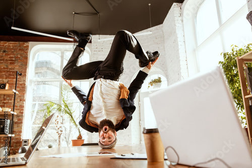 Young caucasian businessman having fun dancing break dance in the modern office at work time with gadgets. Management, freedom, professional occupation, alternative way of working. Loves his job. photo