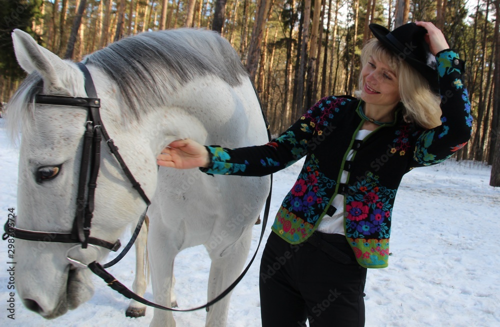Beautiful young blond woman in colorful jacket and black trousers and hat smiles and toches  white horse in bright sunny winter day in the forest 
