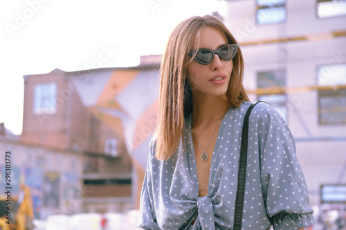 Blonde girl in long blue dress, sunglasses and a small black handbag on her shoulder is walking alone in the city. Fashion and style. Close-up shot. photo