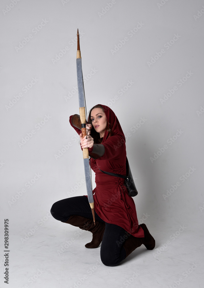 full length portrait of a brunette girl wearing a red fantasy tunic with hood, holding a bow and arrow. Standing pose on a white studio background. full length portrait of a brunette girl wearing a red fantasy tunic with hood, holding a bow and arrow. Standing pose on a white studio background.