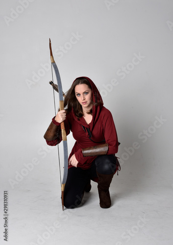 full length portrait of a brunette girl wearing a red fantasy tunic with hood, holding a bow and arrow. Standing pose on a white studio background.