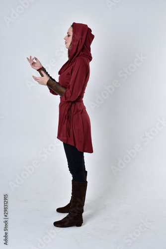 full length portrait of a brunette girl wearing a red fantasy tunic with hood. Standing pose in side profile on a white studio background. photo