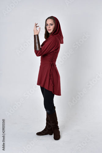 full length portrait of a brunette girl wearing a red fantasy tunic with hood. Standing pose in side profile on a white studio background. photo