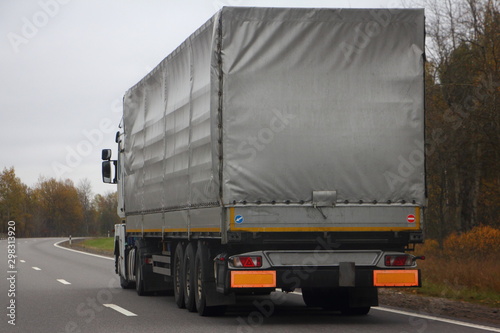 Transport Logistics, international commercial freight by road, gray awning semi truck drive next to the two-lane asphalted country road in the autumn day, side rear view