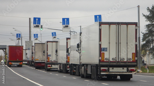 The queue traffic of many trucks with semi-trailers to the control point in the warehouse, inspector in yellow jacket check the cargo documents of the lorry drivers, weight control point