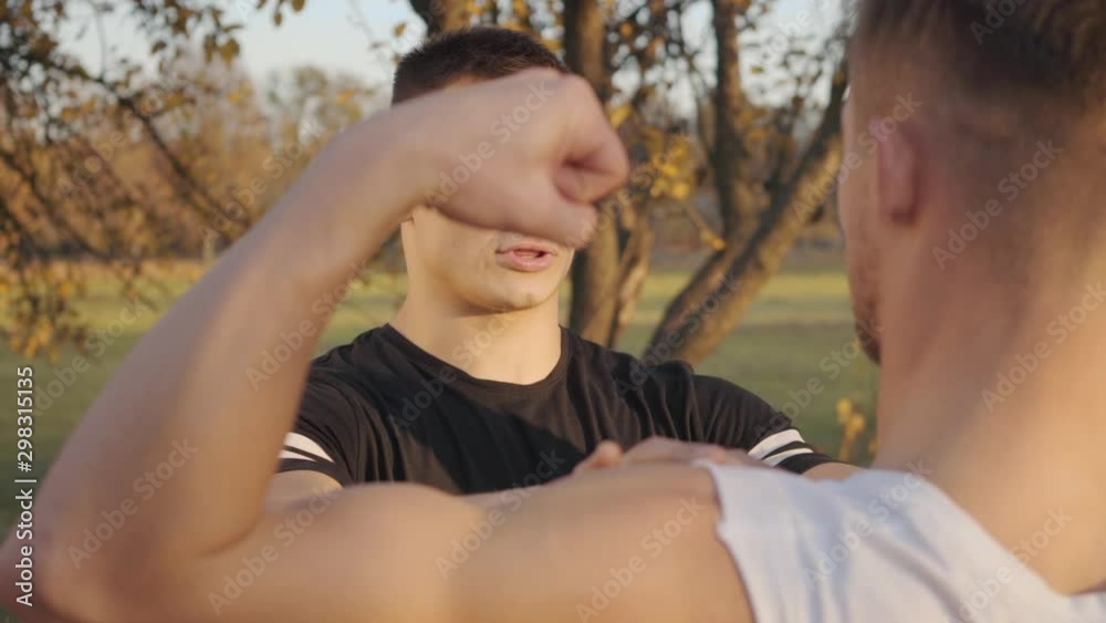 Back view of a young Caucasian sportsman boasting his muscles in sunlight in the autumn park. Young bodybuilder spending sunny day with his trainer outdoors. Healthy lifestyle concept. Back view of a young Caucasian sportsman boasting his muscles in sunlight in the autumn park. Young bodybuilder spending sunny day with his trainer outdoors. Healthy lifestyle concept.