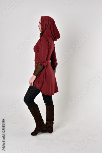 full length portrait of a brunette girl wearing a red fantasy tunic with hood. Standing pose in side profile on a white studio background. photo