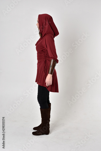 full length portrait of a brunette girl wearing a red fantasy tunic with hood. Standing pose in side profile on a white studio background. photo