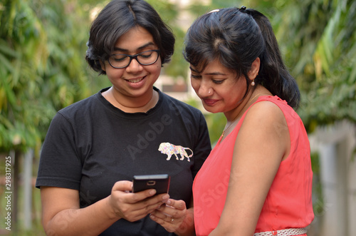 Two friends sitting on a red park bench looking into the mobile phone and smiling and laughing in New Delhi, India © PhotographrIncognito