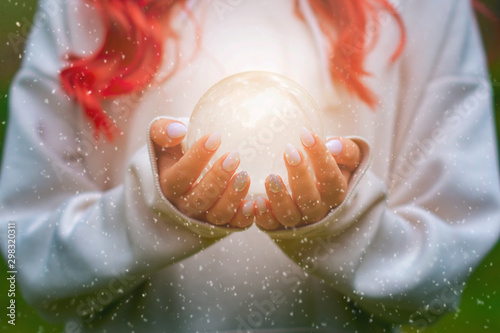 A beautiful young witch girl with long hair is holding a mystical magical, glowing crystal ball in her hands in the forest.