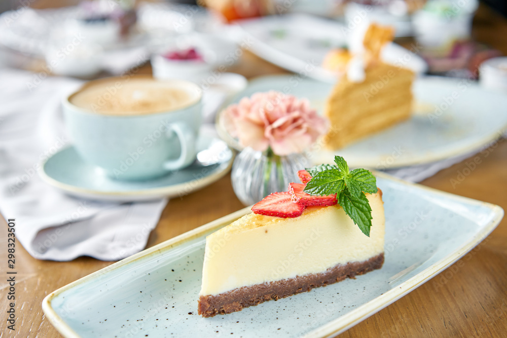 Piece of delicious cheesecake with strawberry and mint leaves on white plate. Breakfast in the cafe, morning coffee. Cappuccino and lots of desserts on the table. - obrazy, fototapety, plakaty 