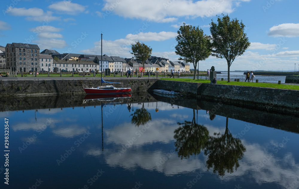 Famous view of the brightly painted houses of Galway city and the River Corrib, with sail boats moored on calm water, with the sky reflecting in it. Taken on a sunny summer day.