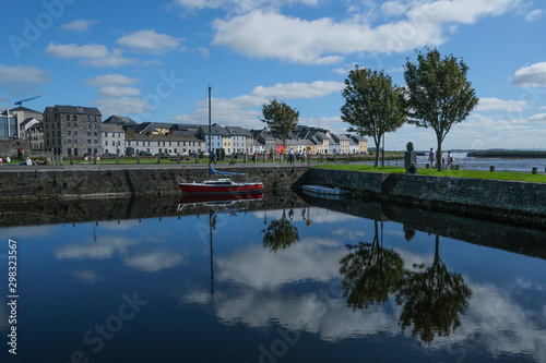 Famous view of the brightly painted houses of Galway city and the River Corrib, with sail boats moored on calm water, with the sky reflecting in it. Taken on a sunny summer day.