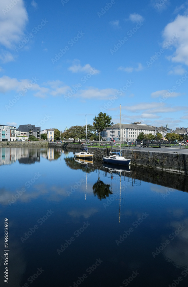 Galway city on a sunny summer day - taken at Eglington Basin near the Spanish Arch showing surrounding buildings & trees, with sailboats moored on calm reflective water.