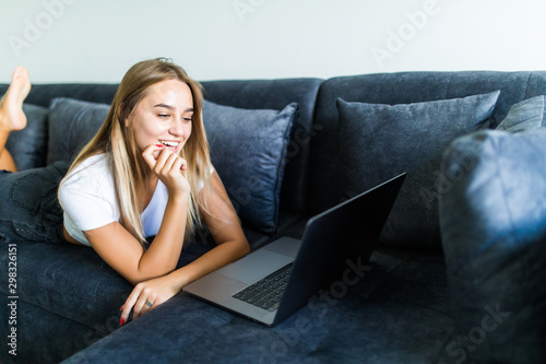 Smiling young woman laying on couch and using laptop at home photo