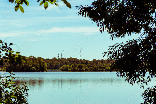Three wind turbines on the horizon behind a forest and a blue lake landscape.