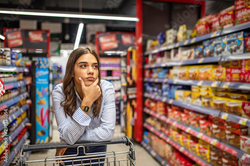 Young woman staying confused in grocery store. Confused woman doesnt know what to buy. Young woman with a shopping cart at supermarket. Looking at supermarket shelf