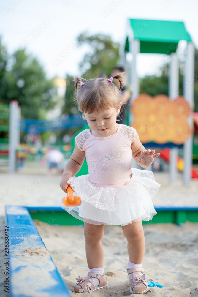 Naklejka premium Cute toddler girl playing in sand on outdoor playground. Beautiful baby in red trousers having fun on sunny warm summer day. Child with colorful sand toys. Healthy active baby outdoors plays games