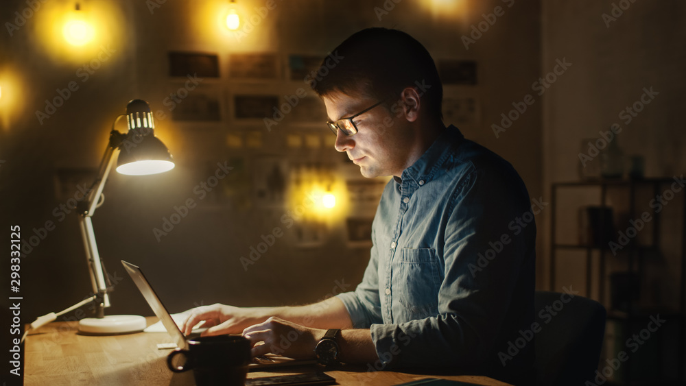 Fototapeta premium Concentrated Man Sitting at His Desk in Office Studio Working on a Laptop in the Evening. Work with Data, Analyzing Statistics.