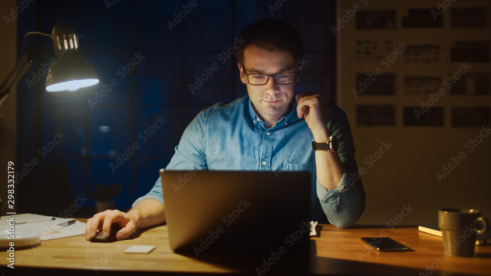 Professional Man Sitting at His Desk in Office Studio Working on a Laptop in the Evening. Man Analyzing Statistics. - obrazy, fototapety, plakaty 