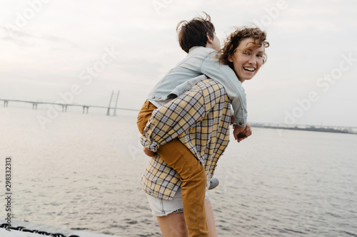 Son enjoying piggyback ride with his young mother while walking. Woman carrying a boy on her back at the outdoors. 