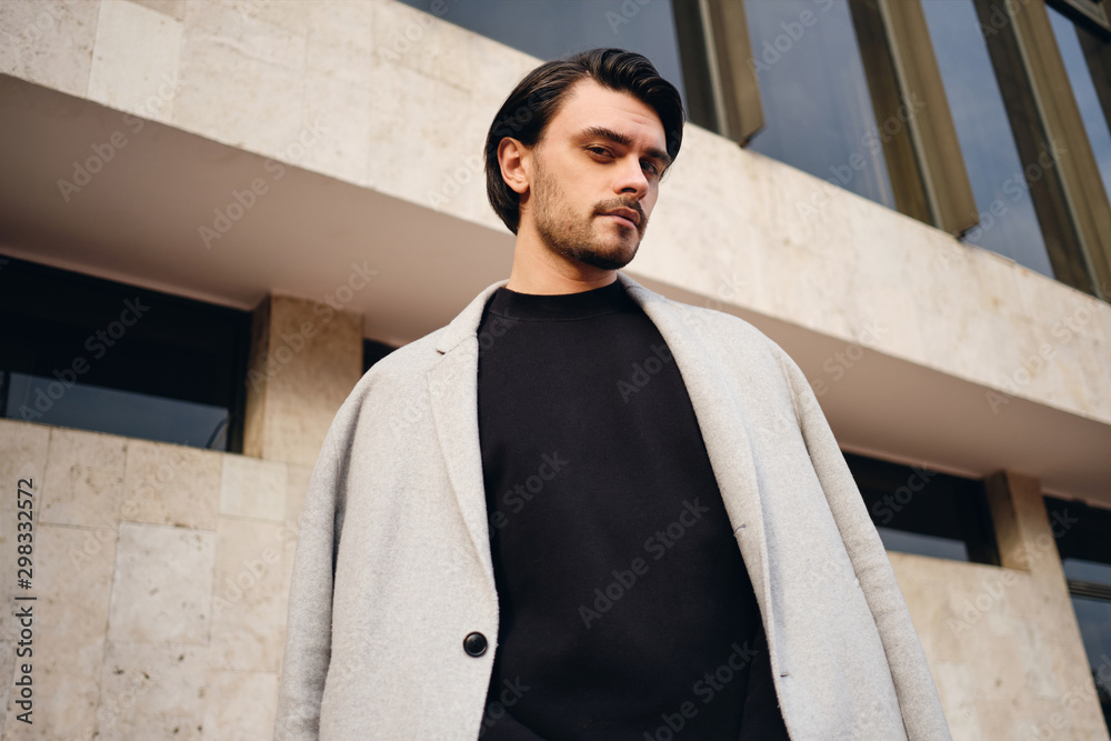 Young attractive stylish man in coat slyly looking in camera outdoor