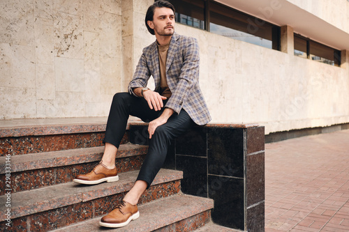 Young handsome stylish man in checkered jacket thoughtfully looking away while sitting outdoor