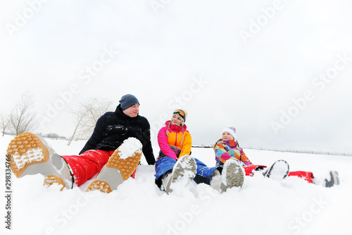 a young family-mom, dad and daughter in bright colored ski suits sitting together in the snow laughing and smiling