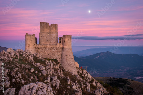 Ruins of medieval castle of Rocca Calascio after sunset and moon rising with mountain landscape and colorful sky in background, Abruzzo, Italy photo