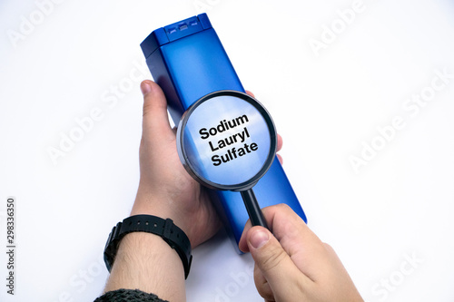 Chemical components on the shampoo label:  Sodium Lauryl Sulfate (sls, sles). A hand holds a blue jar and a magnifier, where the harmful ingredients of a detergent are written in close up. photo