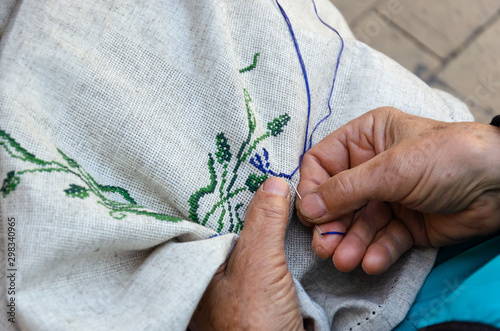 Hands of an elderly woman embroidering a cross-stitch floral pattern on linen fabric. Embroidery, handwork, needlecraft concept. Closeup