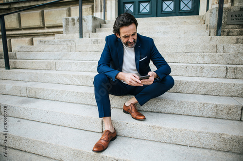 Bearded mature, businessman wearing blue suit sitting on stairs looking at cell phone photo