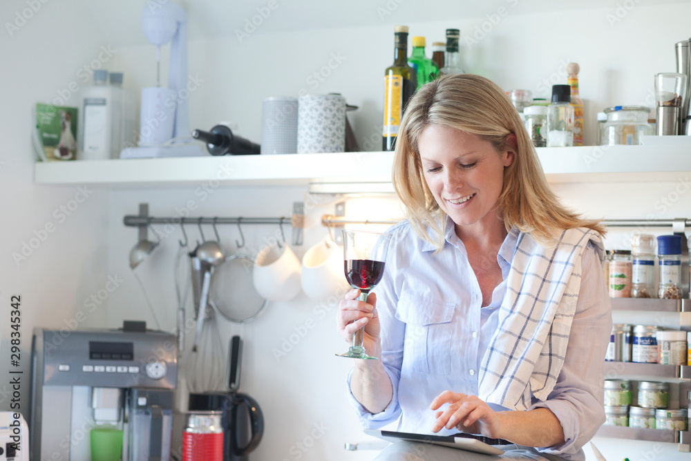 Smiling woman in kitchen drinking glass of red wine and using digital tablet Smiling woman in kitchen drinking glass of red wine and using digital tablet