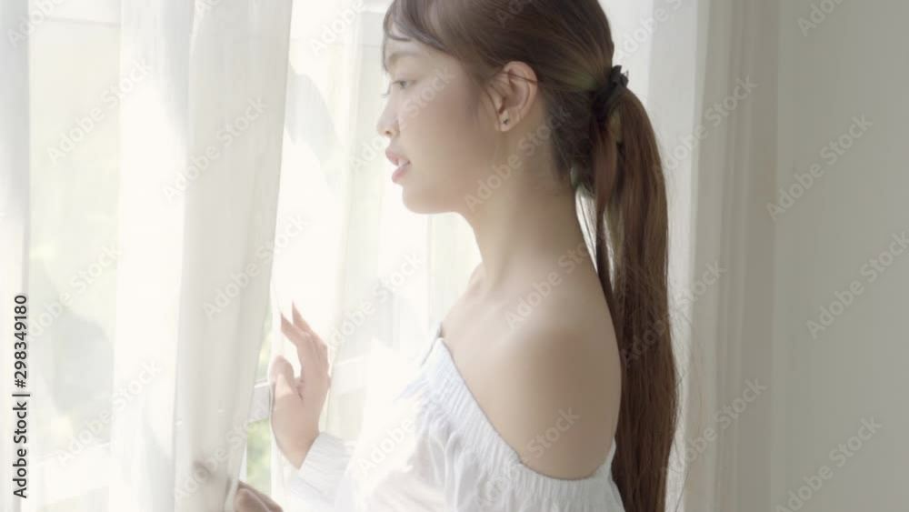 Beautiful portrait young asian woman looking out of window in bedroom at morning after wake up, girl relax with smiling and happy, lifestyle concept, slow motion. Beautiful portrait young asian woman looking out of window in bedroom at morning after wake up, girl relax with smiling and happy, lifestyle concept, slow motion.