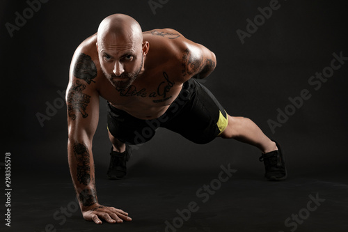 Athletic bald, tattooed man in black shorts and sneakers is posing against a black background. Close-up portrait.