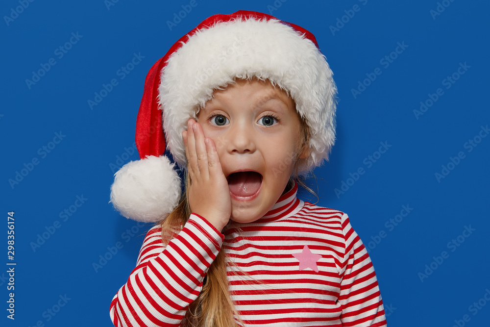 Portrait of surprised and shocked little toddler child girl in Santa hat standing isolated over blue background. Looking at camera. hands near open mouth. Happy New Year and Christmas holiday concept Portrait of surprised and shocked little toddler child girl in Santa hat standing isolated over blue background. Looking at camera. hands near open mouth. Happy New Year and Christmas holiday concept