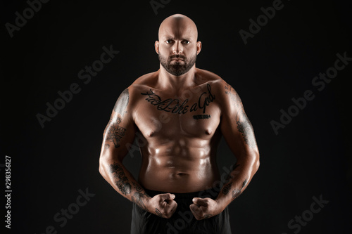 Athletic bald, bearded, tattooed man in black shorts is posing against a black background. Close-up portrait.