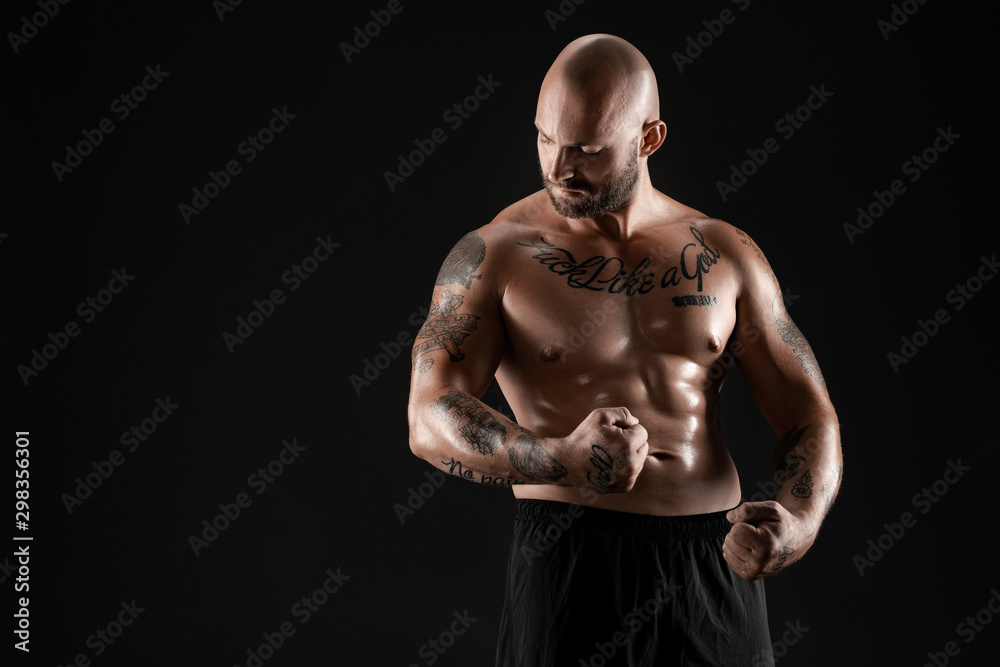 Fototapeta premium Athletic bald, bearded, tattooed man in black shorts is posing against a black background. Close-up portrait.