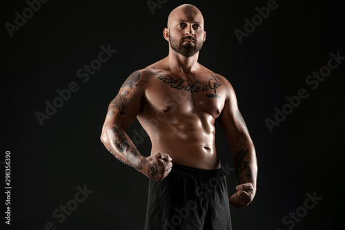 Athletic bald, bearded, tattooed man in black shorts is posing against a black background. Close-up portrait. © nazarovsergey