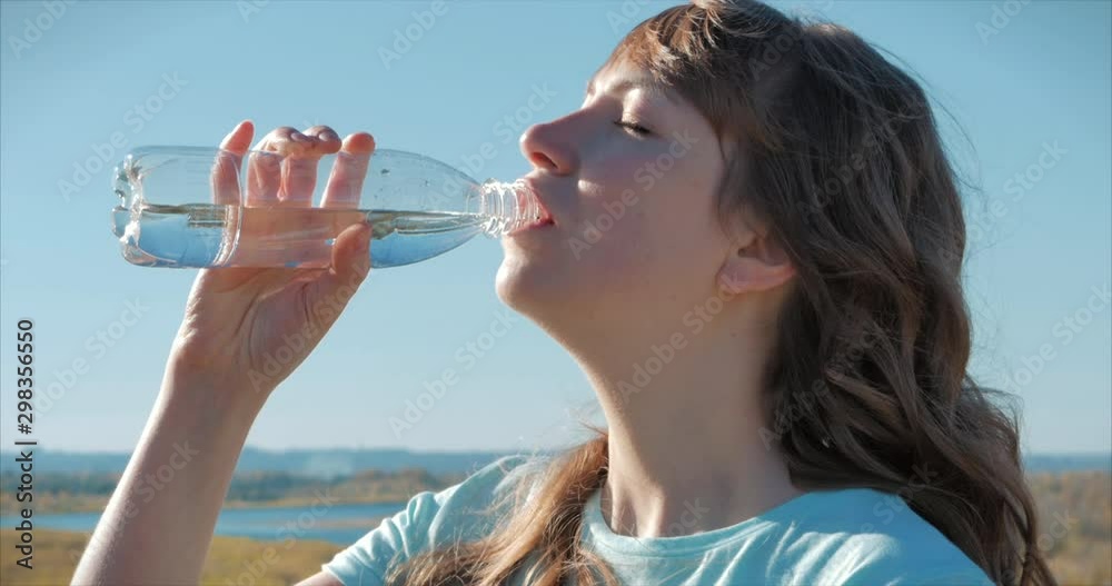 Woman of European Appearance in the Open air Drinks Water from a Bottle Against a Clear Blue Sky. Healthy Lifestyle. Woman of European Appearance in the Open air Drinks Water from a Bottle Against a Clear Blue Sky. Healthy Lifestyle.