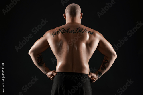 Athletic bald, bearded, tattooed man in black shorts is posing against a black background. Close-up portrait. © nazarovsergey