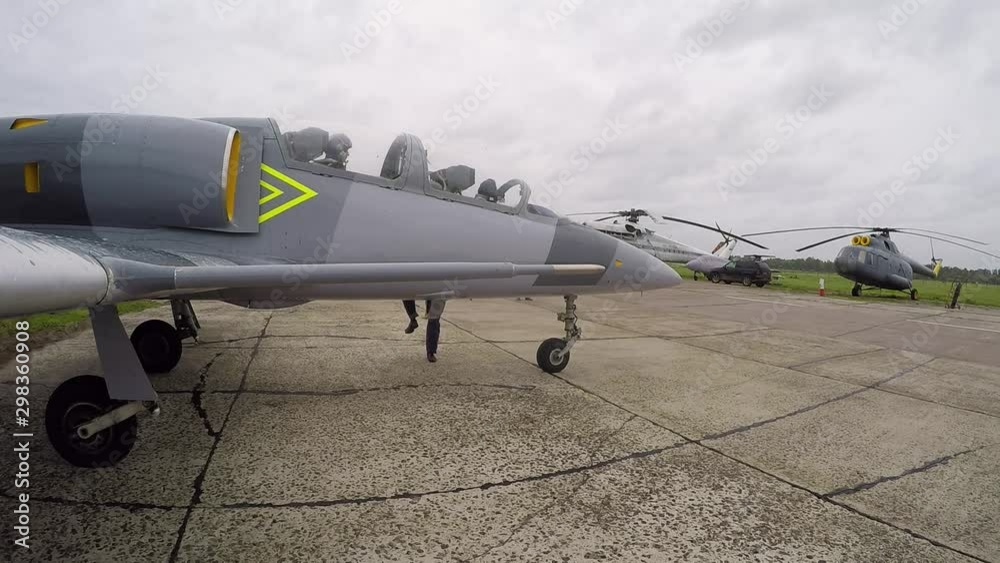 The assistant pilot close the cockpit. The crew of the gray fighter training aircraft is ready for take-off. View from the right wing on the background of helicopters on green grass on a cloudy day. The assistant pilot close the cockpit. The crew of the gray fighter training aircraft is ready for take-off. View from the right wing on the background of helicopters on green grass on a cloudy day.
