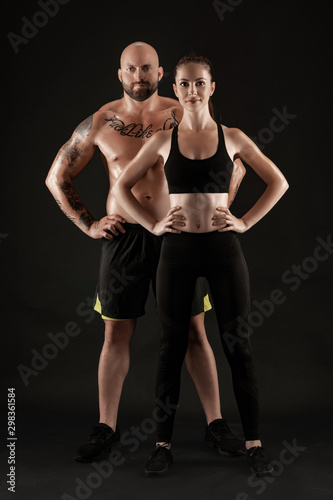 Athletic man in shorts and sneakers with brunette woman in leggings and top posing on black background. Fitness couple, gym concept.