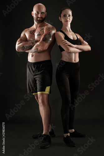 Athletic man in shorts and sneakers with brunette woman in leggings and top posing on black background. Fitness couple, gym concept.